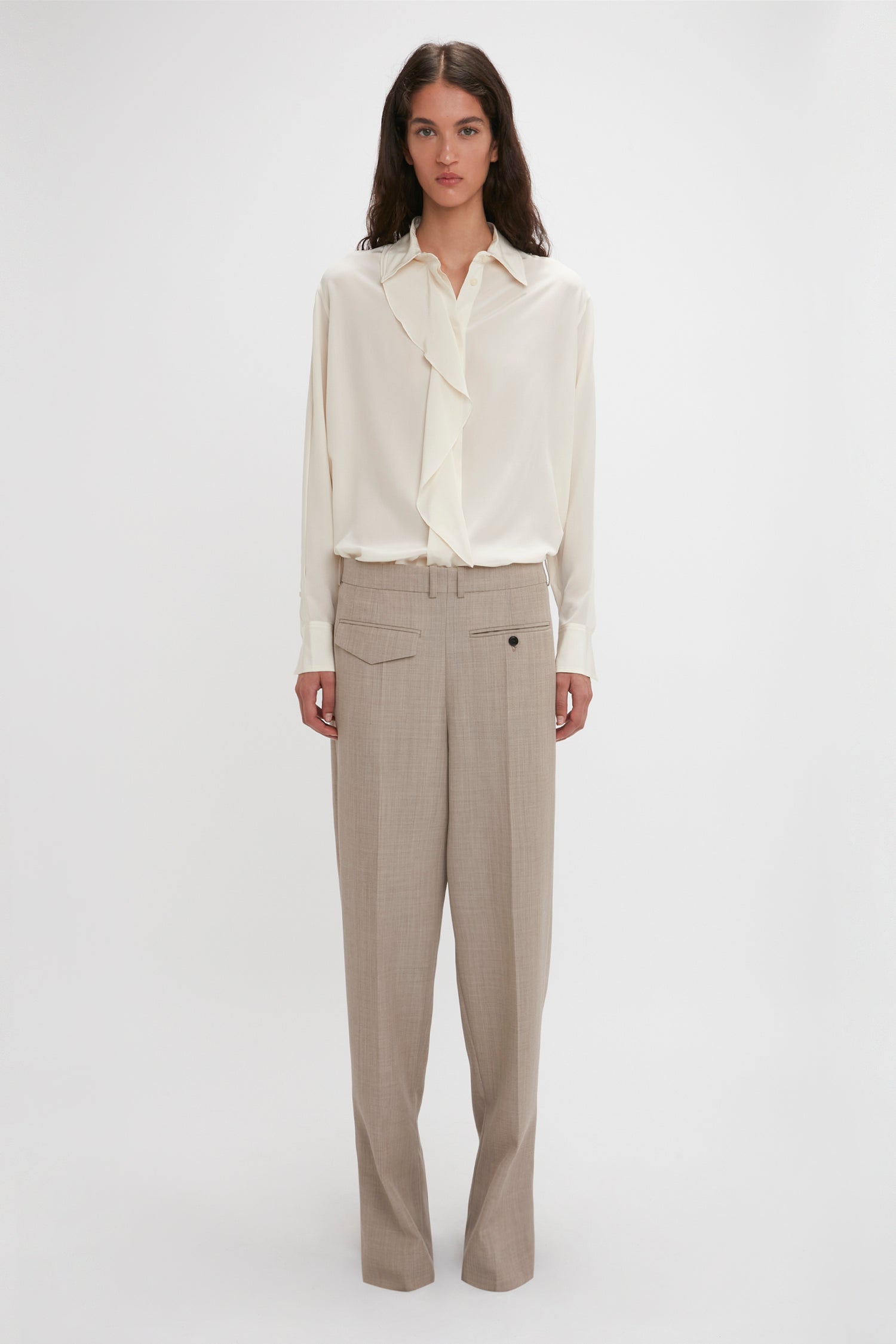 A person stands against a plain background, wearing an Asymmetric Ruffle Blouse In Ivory by Victoria Beckham with ruffle detail and voluminous sleeves, paired with beige wide-leg trousers.