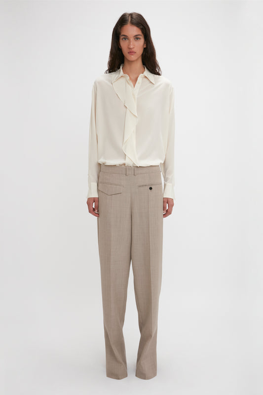 A person stands against a plain background, wearing an Asymmetric Ruffle Blouse In Ivory by Victoria Beckham with ruffle detail and voluminous sleeves, paired with beige wide-leg trousers.