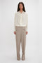 A person stands against a plain background, wearing an Asymmetric Ruffle Blouse In Ivory by Victoria Beckham with ruffle detail and voluminous sleeves, paired with beige wide-leg trousers.