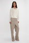 A person with long dark hair is wearing an Asymmetric Ruffle Blouse In Ivory by Victoria Beckham, featuring voluminous sleeves and tan wide-leg trousers, standing against a plain white backdrop.