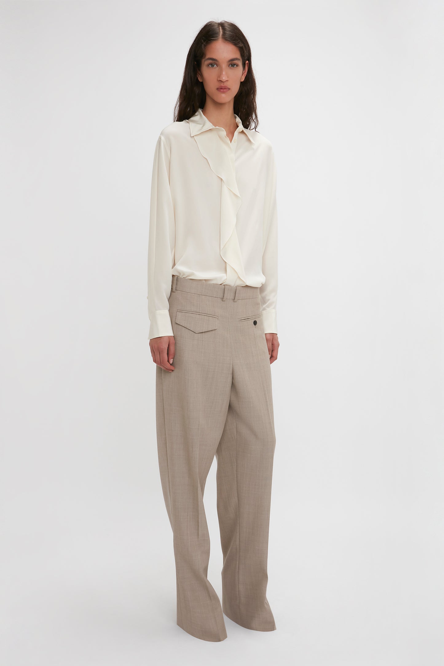 A woman with long hair is wearing the Victoria Beckham Asymmetric Ruffle Blouse In Ivory and beige wide-leg trousers. She is standing against a plain white background.