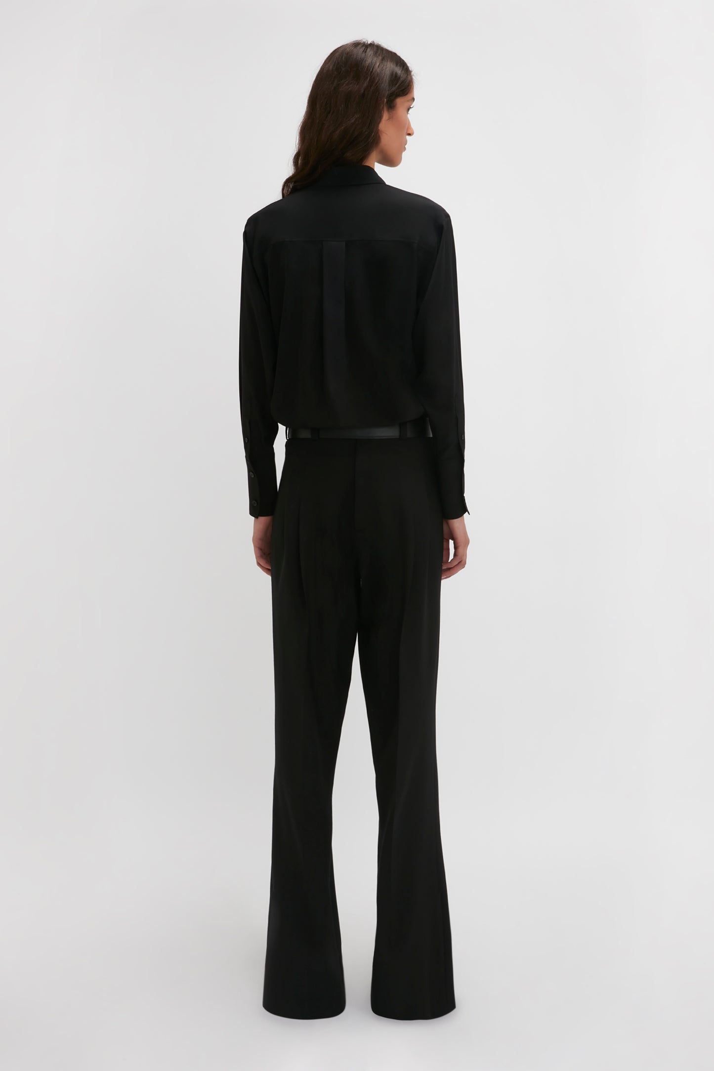 A person with long hair is standing with their back to the camera, wearing a black Victoria Beckham Contrast Bib Shirt In Black and black pants against a plain white background.