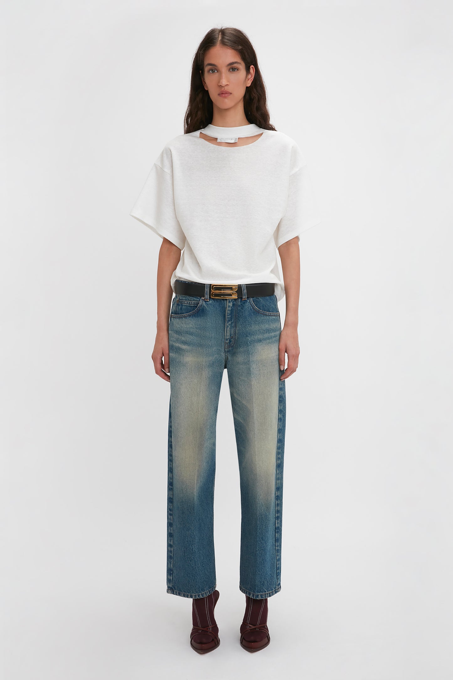 A woman stands against a plain white background, wearing a white, relaxed cut Victoria Beckham Bonded Jersey Tee In White, blue jeans with a slightly faded effect, a black belt with a gold buckle, and dark brown ankle boots. This ensemble exudes modern nonchalance.