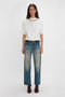 A woman stands against a plain white background, wearing a white, relaxed cut Victoria Beckham Bonded Jersey Tee In White, blue jeans with a slightly faded effect, a black belt with a gold buckle, and dark brown ankle boots. This ensemble exudes modern nonchalance.