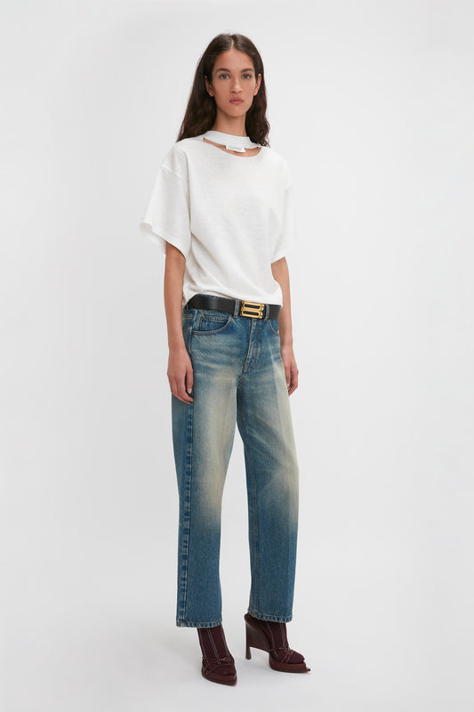 A person with long hair is wearing a white Bonded Jersey Tee In White by Victoria Beckham with a unique neckline, blue jeans, a black belt, and dark red high-heeled shoes, exuding an air of modern nonchalance while standing against a plain white background.