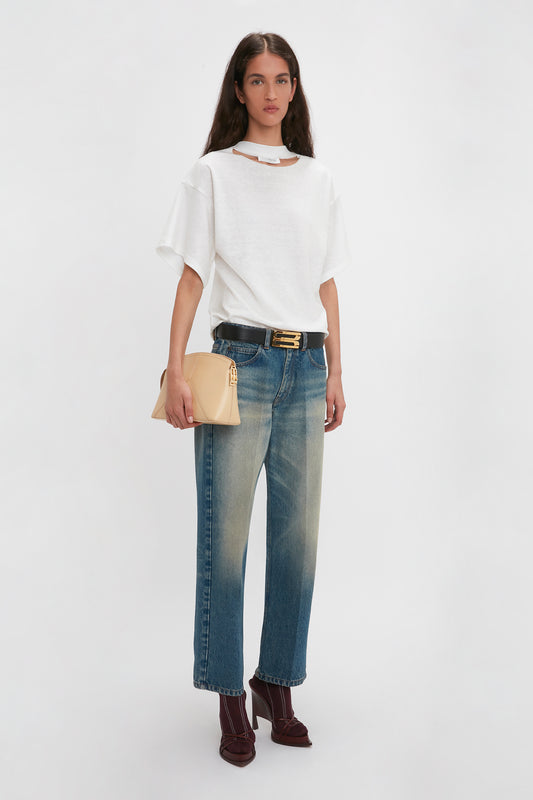 A person stands against a white background, exuding modern nonchalance in a relaxed cut Bonded Jersey Tee In White by Victoria Beckham, faded jeans, burgundy shoes, and holding a beige clutch.