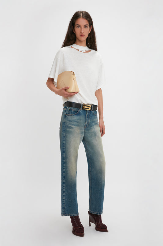 A woman stands against a plain background, showcasing modern nonchalance in a Victoria Beckham Bonded Jersey Tee In White, blue jeans, and burgundy high heels. She completes her relaxed cut outfit with a beige handbag tucked under her arm.