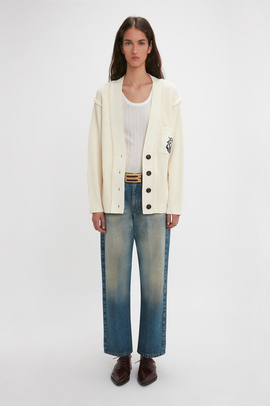 A person stands against a plain background wearing a Victoria Beckham Relaxed Fit Cardigan In Natural over a white top, blue jeans, and dark brown shoes.