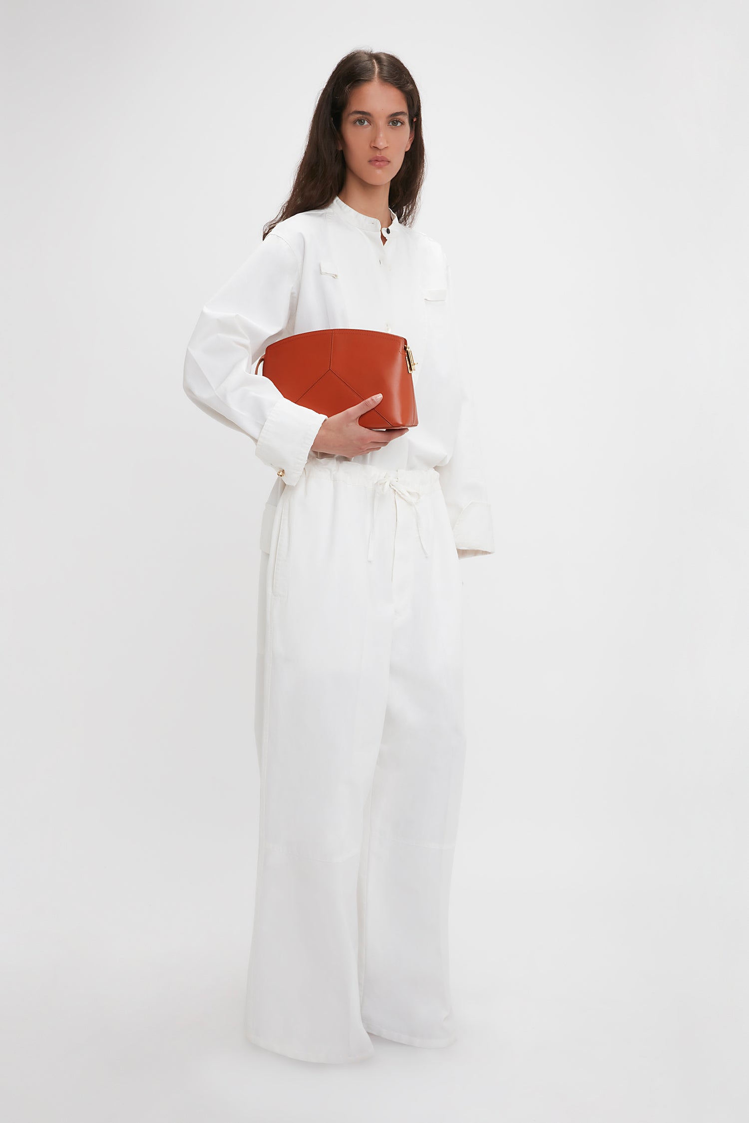 A person stands against a plain background, dressed entirely in white and holding a large, geometric Victoria Clutch Bag In Tan Leather by Victoria Beckham.