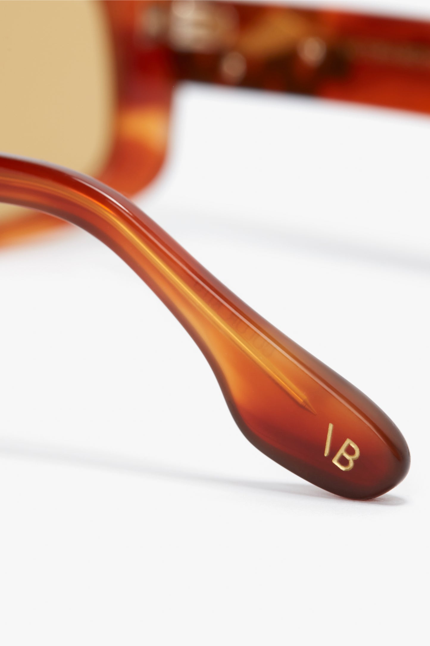 Close-up of the arm of a pair of Chain Detail Rectangular Frame Sunglasses In Striped Blonde Havana by Victoria Beckham with a chain detail and the initials "IB" engraved on the side. The background is blurred.