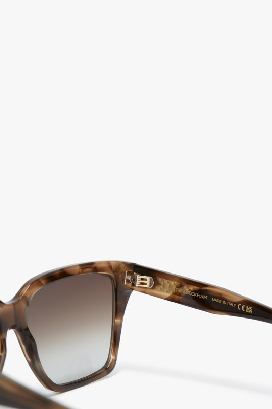 Close-up of the inside arm of a pair of Victoria Beckham Soft Square Frame Sunglasses In Striped Khaki Havana, showing manufacturer details, including "MADE IN ITALY" and certification marks. The frame is brown tortoiseshell with gradient lenses in a contemporary design.