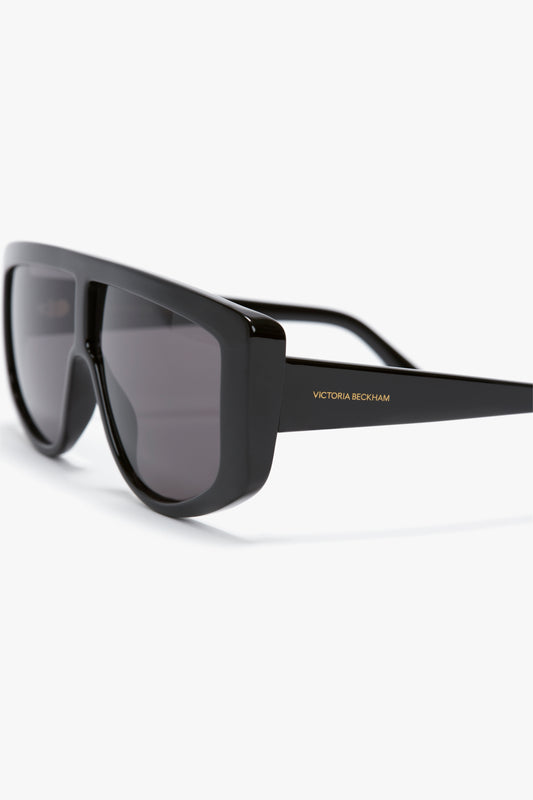 Close-up of black oversized rectangular sunglasses with dark lenses, featuring "Victoria Beckham" written in gold on the temple. The product is Acetate Visor Sunglasses In Black by Victoria Beckham.