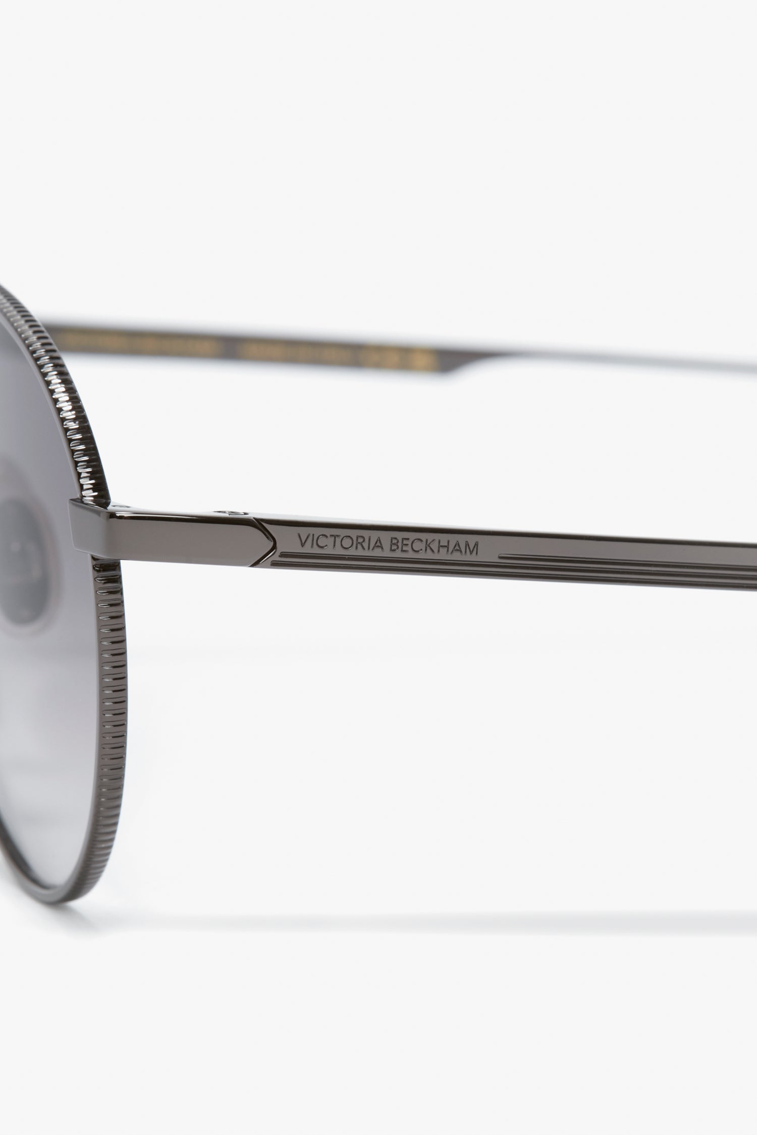 Close-up of a pair of V Metal Pilot Sunglasses In Grey Gradient with the "Victoria Beckham" logo inscribed on the metal arm. The sleek frame sports a gunmetal grey gradient design, dark lenses, and a textured edge.