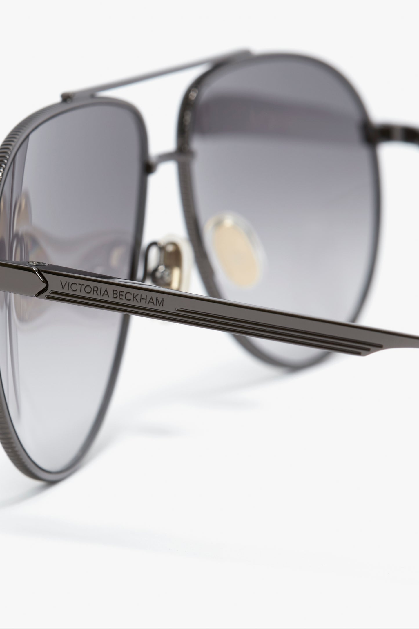 Close-up of sleek, metal-framed V Metal Pilot Sunglasses In Grey Gradient with a gunmetal grey gradient design and "Victoria Beckham" inscribed on the arm. The lenses are shaded, and the image focuses on the brand's logo.