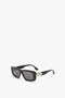 A pair of Victoria Beckham Chain Detail Rectangular Frame Sunglasses In Black with dark lenses is shown on a white background. The arms feature a metallic, interlocking square design near the hinges, echoing modern eyewear trends with a Chain Detail Rectangular Frame look.