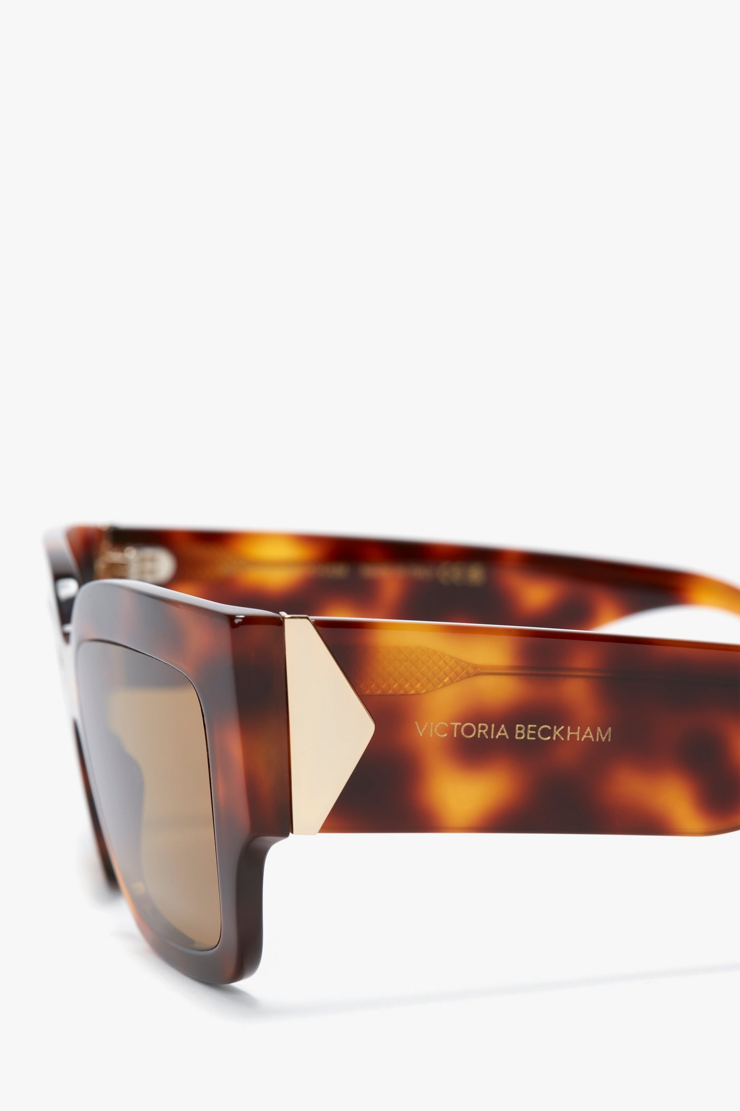 Close-up of V Plaque Rectangular Frame Sunglasses In Tortoise with a gold triangular detail on the rectangular frame and the text "Victoria Beckham" inscribed on the arm.