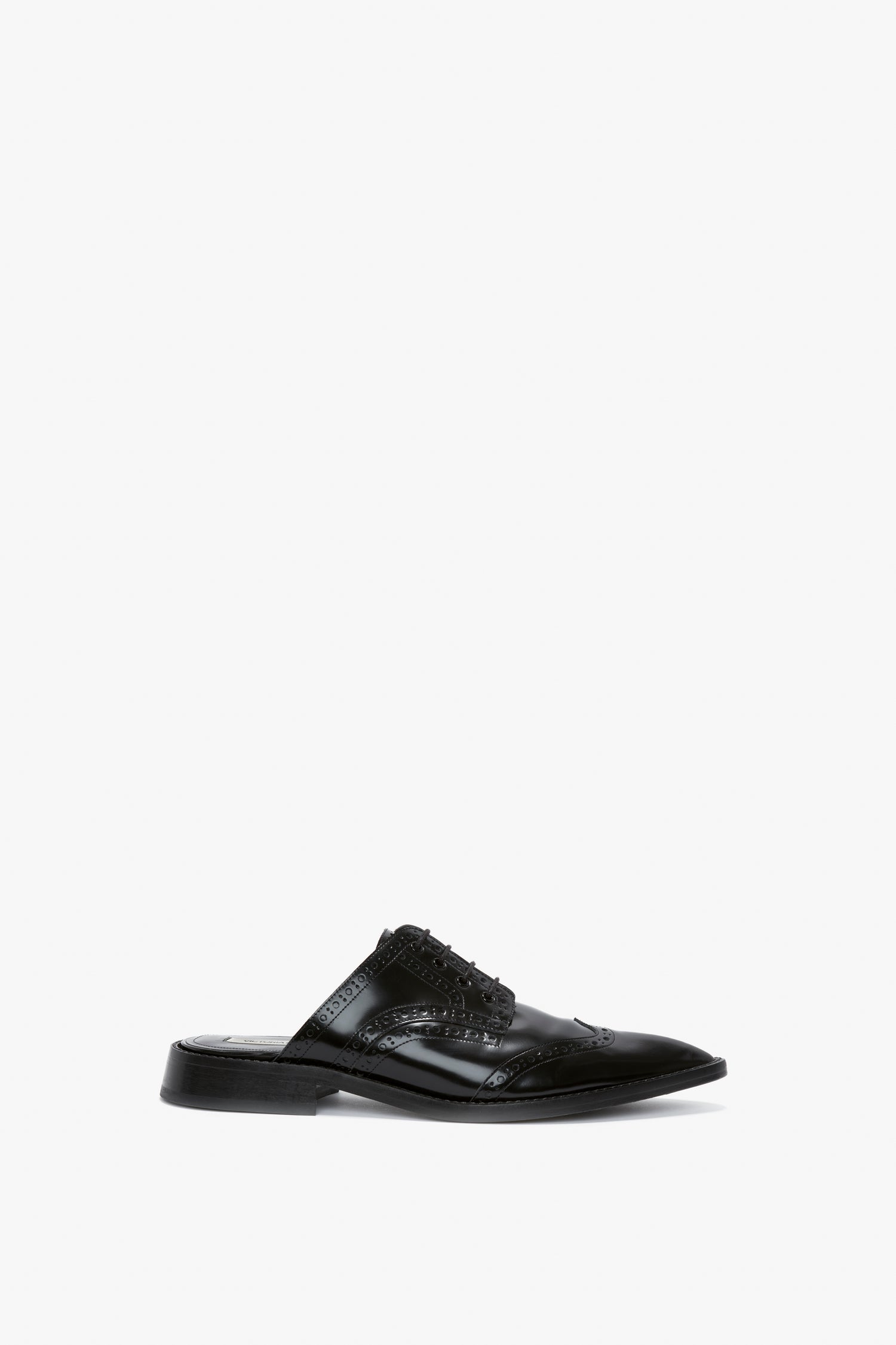 A black leather slip-on shoe, the Flat Lace Up Mules In Black Leather by Victoria Beckham, featuring wingtip brogue detailing and a low heel, crafted from luxurious calfskin with a pointy toe silhouette, set against a white background.