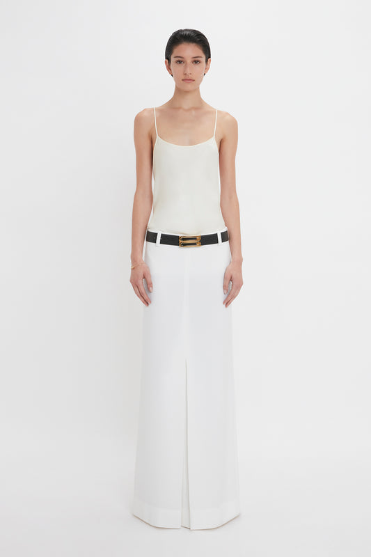 A person stands facing forward, wearing a white sleeveless top, a black belt, and an Exclusive Tailored Floor-Length Pleated Skirt In Ivory by Victoria Beckham. The background is plain white.