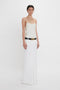 A person in a white sleeveless top and long Exclusive Tailored Floor-Length Pleated Skirt In Ivory by Victoria Beckham stands against a white background. The pleated skirt has a black belt with a gold buckle.