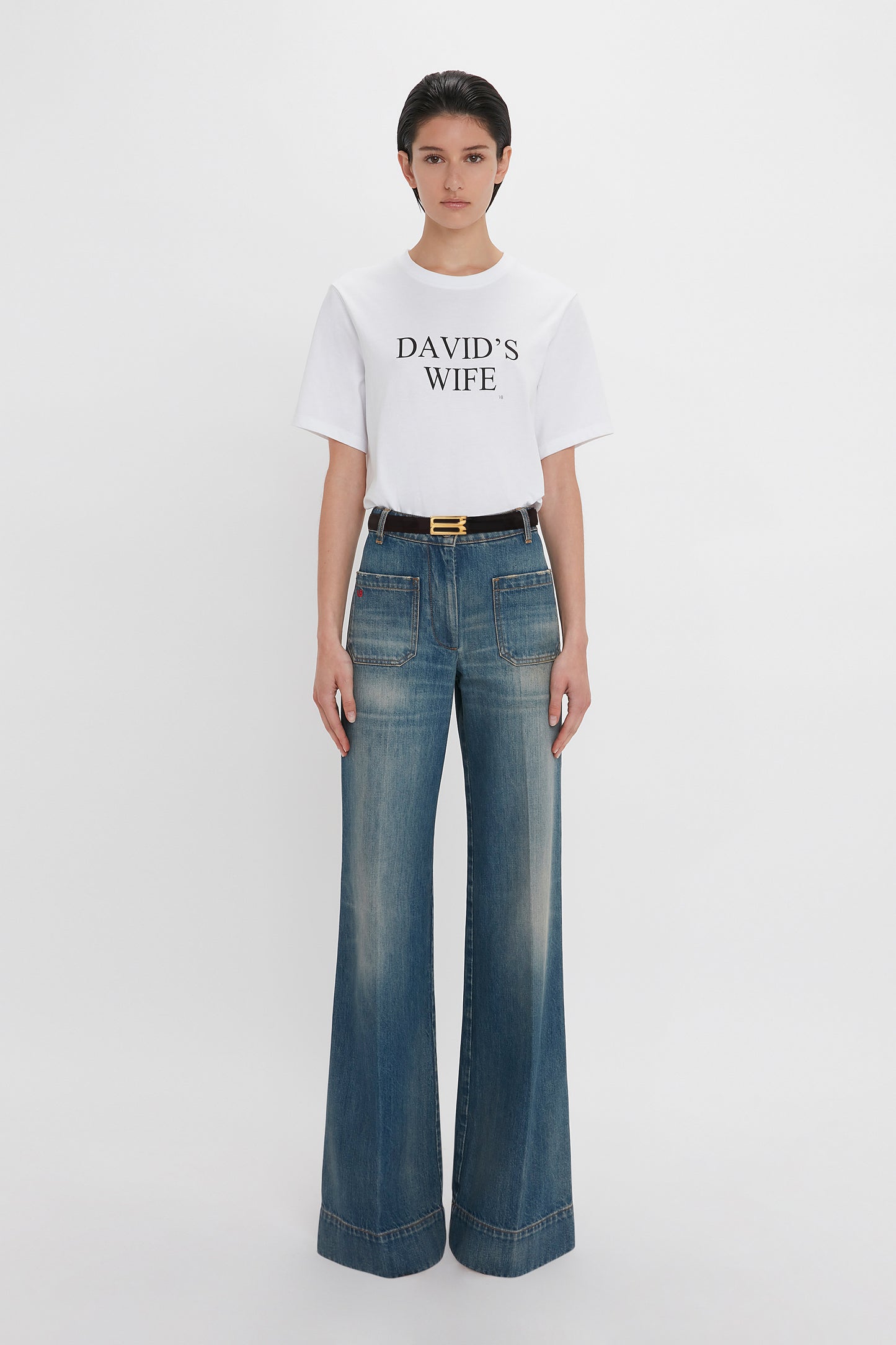 A person stands against a white background wearing a white T-shirt with the text "DAVID'S WIFE" and Victoria Beckham's Alina High Waisted Jean In Indigrey Wash featuring seventies-style pockets.