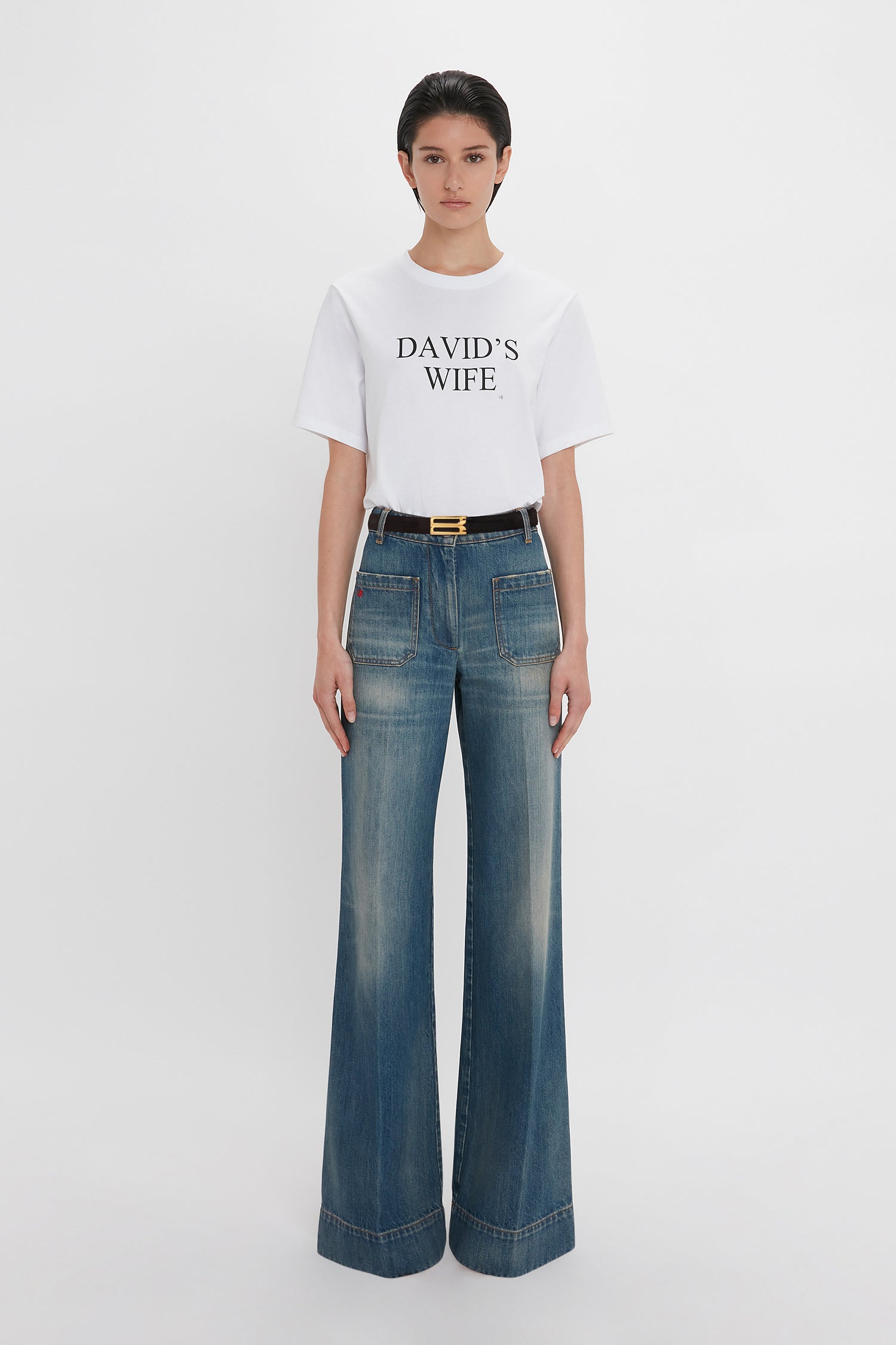 A person stands against a white background wearing a white T-shirt with the text "DAVID'S WIFE" and Victoria Beckham's Alina High Waisted Jean In Indigrey Wash featuring seventies-style pockets.