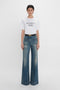 A person stands against a white background wearing a white T-shirt with the text "DAVID'S WIFE" and Victoria Beckham's Alina High Waisted Jean In Indigrey Wash featuring seventies-style pockets.