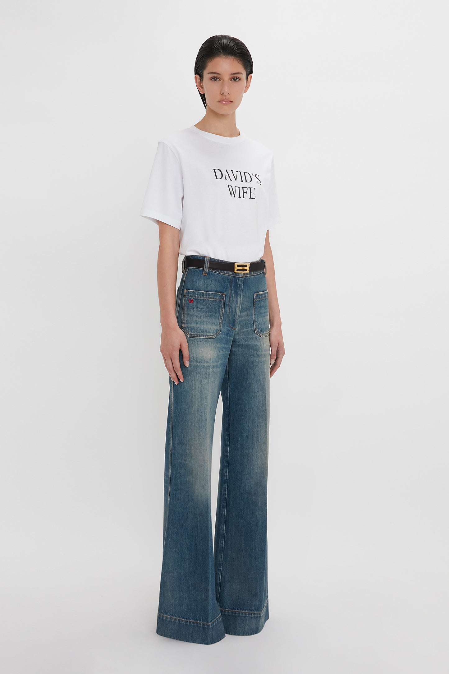 Person wearing a white T-shirt with "DAVID'S WIFE" text, a dark belt, and Victoria Beckham Alina High Waisted Jean In Indigrey Wash with seventies-style pockets, standing against a plain white background.