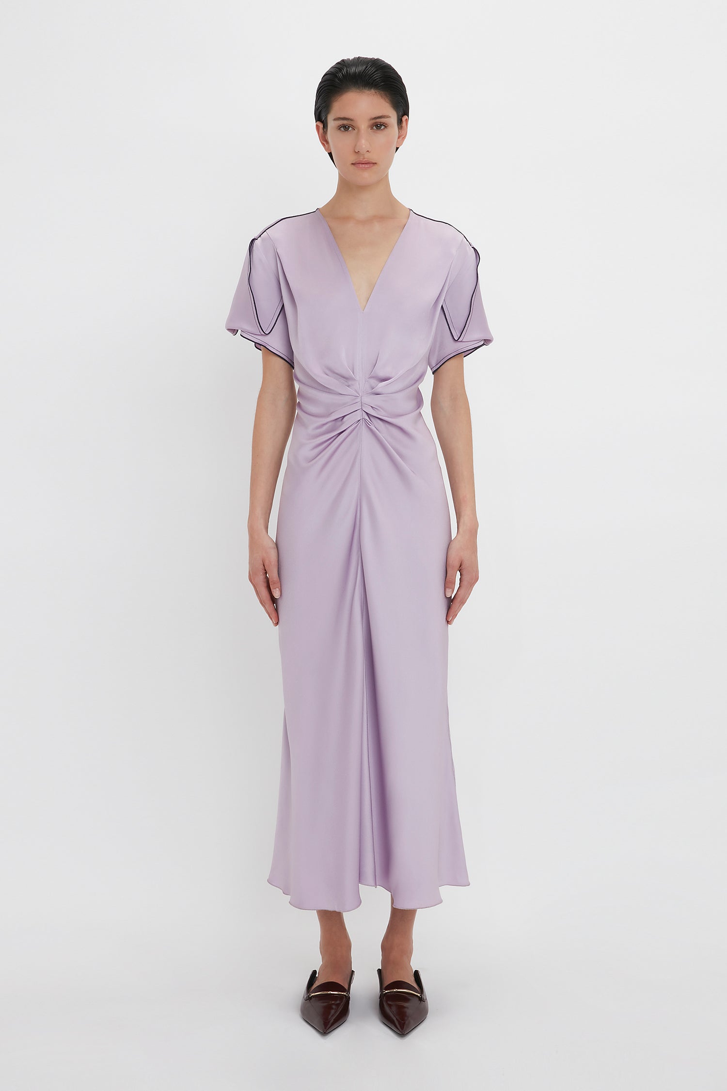 Person wearing the Gathered V-Neck Midi Dress In Petunia by Victoria Beckham with short sleeves and a waist-defining pleat, paired with pointed brown shoes. They are standing against a plain white background.