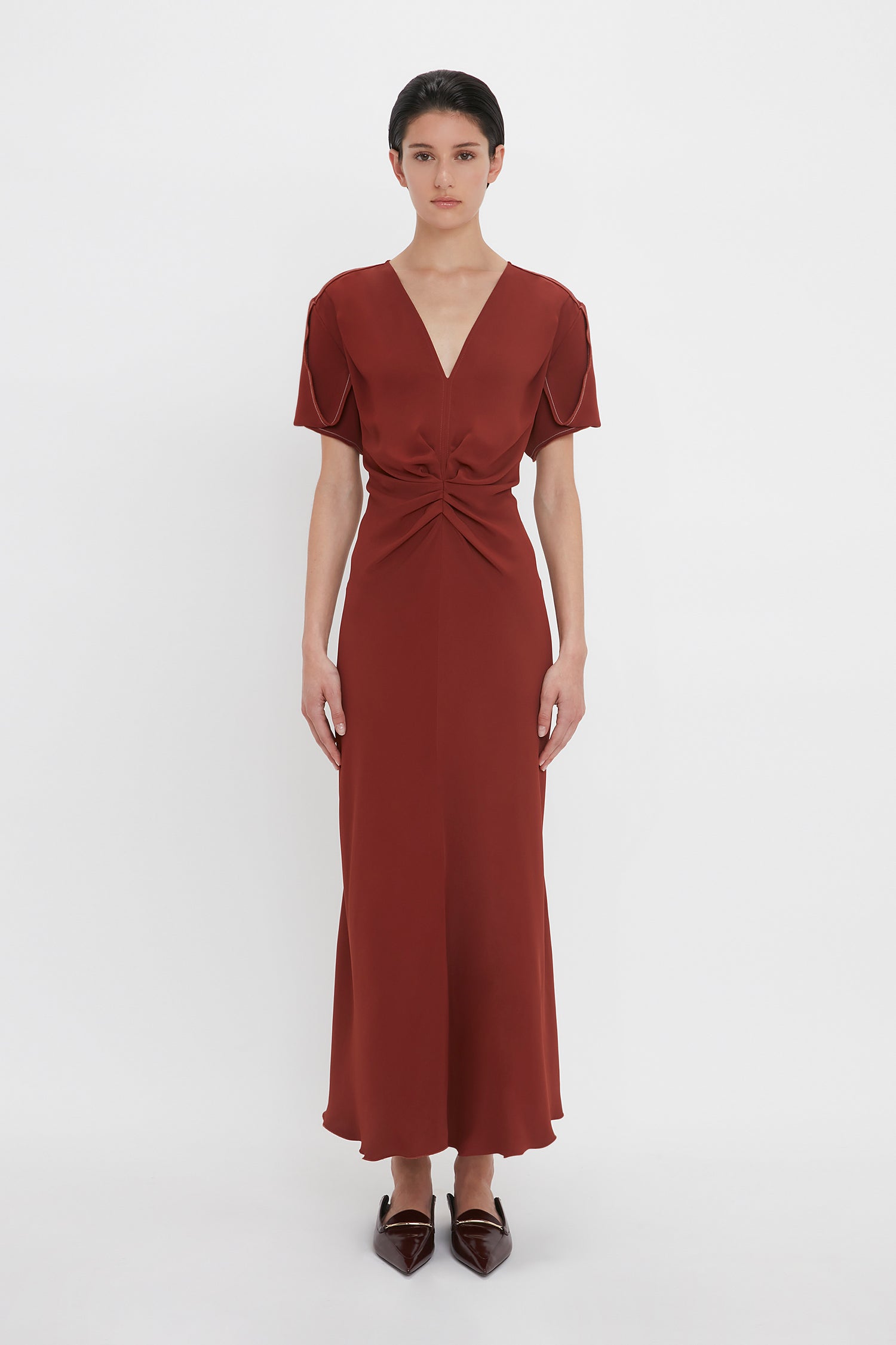 A person stands against a white background wearing a Gathered V-Neck Midi Dress In Russet by Victoria Beckham. The short-sleeved dress, made from figure-flattering stretch fabric, features a waist-defining pleat detail and knotted accent. The individual has short, dark hair and pointed shoes.