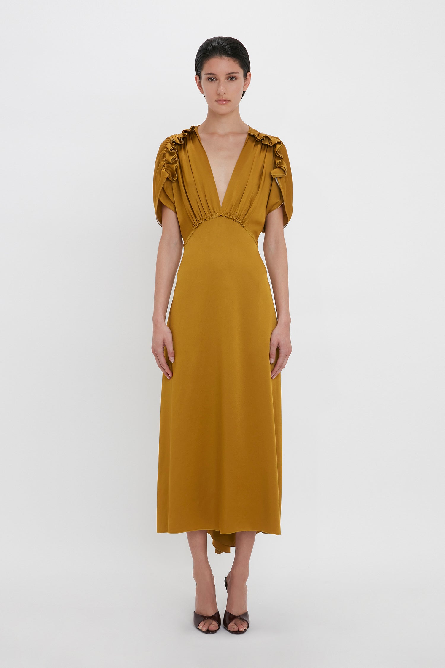 A person stands in a Victoria Beckham V-Neck Ruffle Midi Dress In Harvest Gold, its seductive quality highlighted by the deep V-neckline, short ruched sleeves, and mid-calf length hemline. They wear black heels against a plain white background.