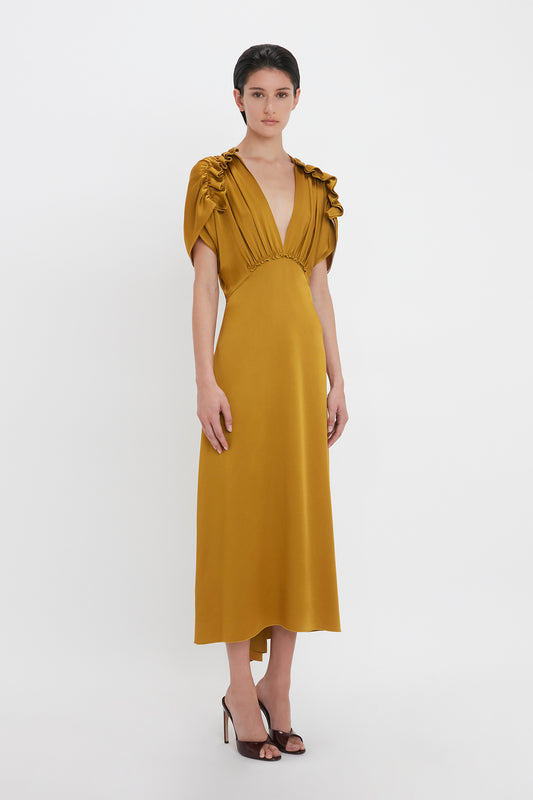 A person wearing a Victoria Beckham V-Neck Ruffle Midi Dress In Harvest Gold with short ruffled sleeves and black high-heeled shoes stands against a plain white background, exuding a seductive quality.