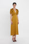 A person wearing a Victoria Beckham V-Neck Ruffle Midi Dress In Harvest Gold with short ruffled sleeves and black high-heeled shoes stands against a plain white background, exuding a seductive quality.