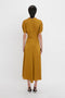 A person is standing with their back to the camera, wearing a Victoria Beckham V-Neck Ruffle Midi Dress In Harvest Gold and black high-heeled shoes that lend a subtle, seductive quality to their look.