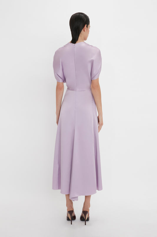 A person is standing facing away from the camera, wearing a Victoria Beckham V-Neck Ruffle Midi Dress In Petunia and black high-heeled shoes. The background is plain white, adding to the elegance reminiscent of Victoria Beckham's timeless style.