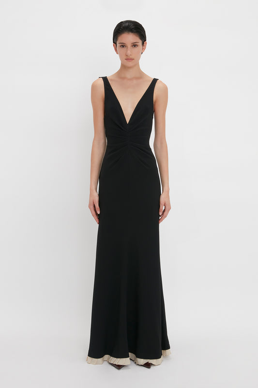 A person stands against a plain background wearing a Victoria Beckham V-Neck Gathered Waist Floor-Length Gown In Black with a deep V-neckline, slightly flared at the hem, creating a flattering silhouette.