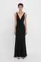 A person stands against a plain background wearing a Victoria Beckham V-Neck Gathered Waist Floor-Length Gown In Black with a deep V-neckline, slightly flared at the hem, creating a flattering silhouette.