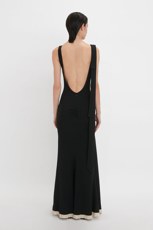 Person wearing a black, backless V-Neck Gathered Waist Floor-Length Gown In Black by Victoria Beckham with white trim at the hem, featuring a deep-V neckline and a flattering silhouette, standing against a plain white background.