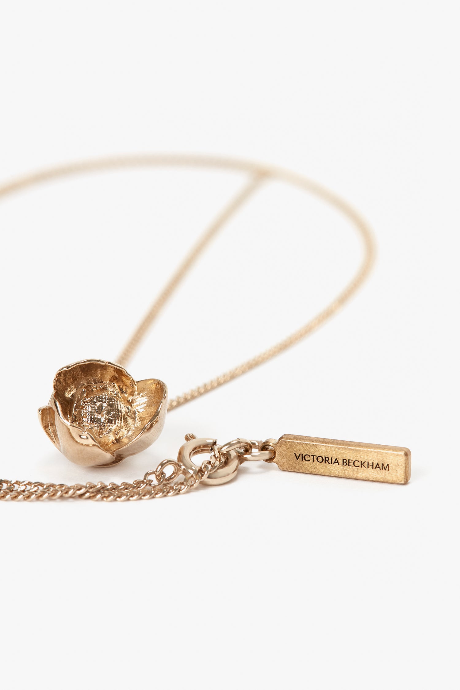 Close-up of an Exclusive Camellia Flower Necklace In Gold featuring a textured pendant and a rectangular tag inscribed with "Victoria Beckham." Expertly handcrafted, the gold brushed brass piece is laid out on a white surface.