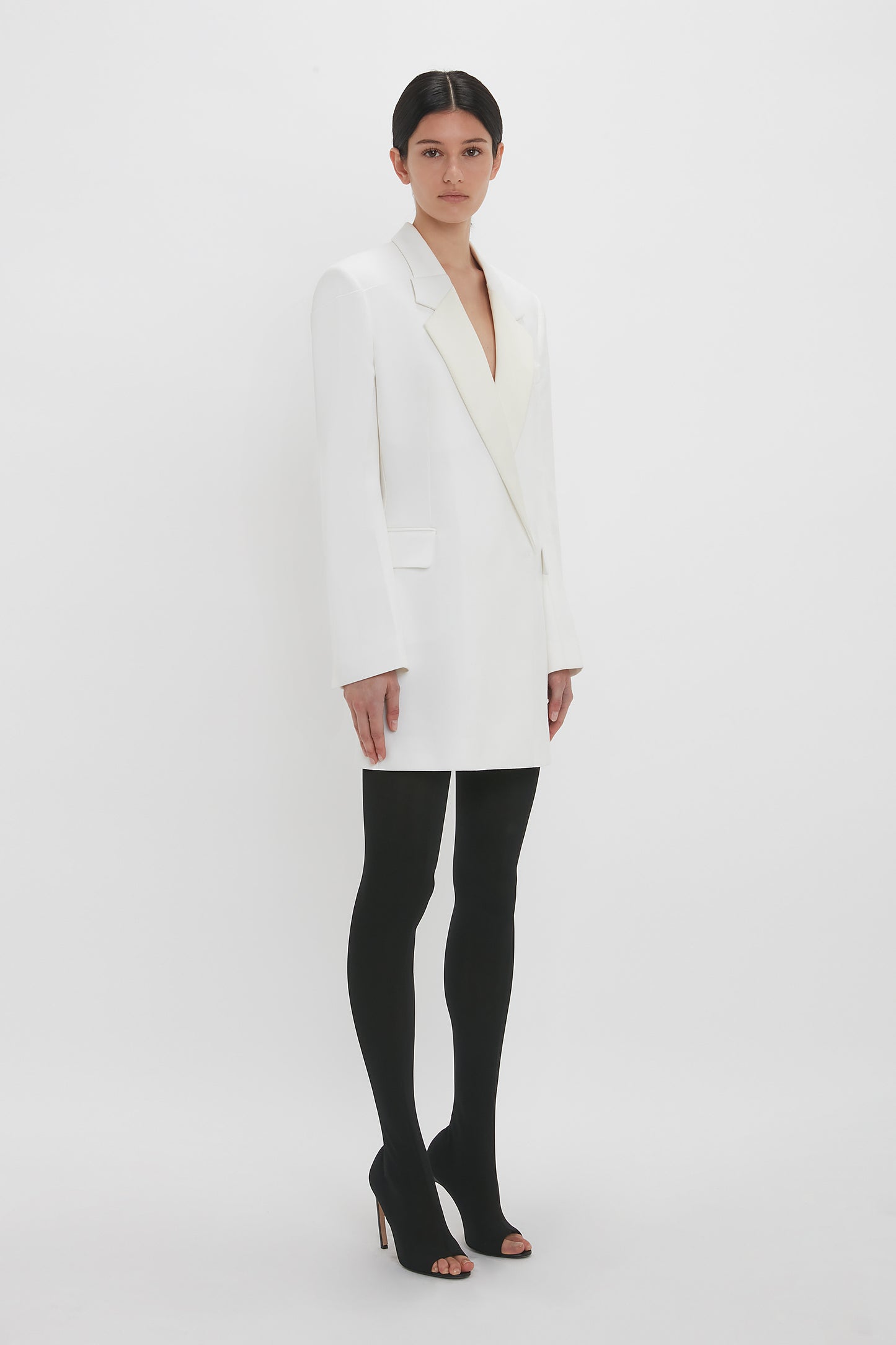 A woman dressed in a stylish double-breasted Exclusive Fold Shoulder Detail Dress In Ivory by Victoria Beckham and black tights stands against a white background, looking directly at the camera.