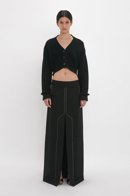 A person stands against a plain white background, wearing a black, three-button cropped cardigan paired with a matching Deconstructed Floor-Length Skirt In Black by Victoria Beckham. The skirt features a front slit, visible stitching, and an elongating silhouette that enhances the overall look.