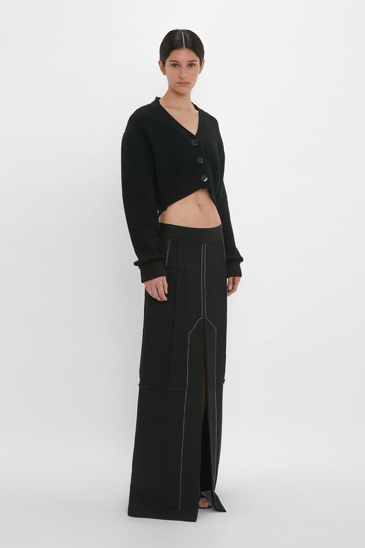 A person stands in a black cardigan and the Victoria Beckham Deconstructed Floor-Length Skirt In Black. The cardigan is cropped, and the floor-grazing skirt features a front slit, creating an elongating silhouette against the white background.