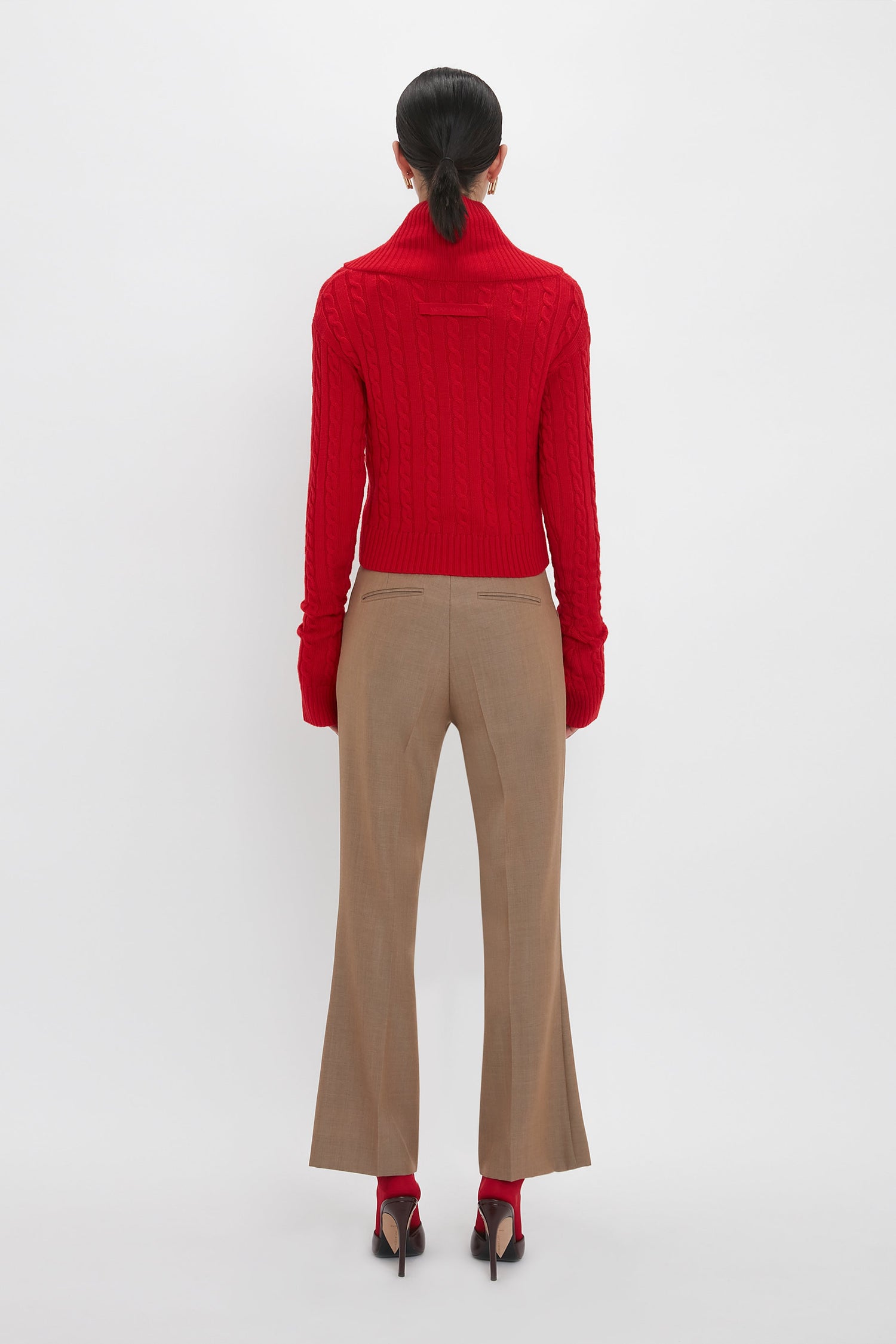 Person standing against a plain background, facing away. They are wearing a red turtleneck sweater, Wide Cropped Flare Trouser In Tobacco by Victoria Beckham that offer a flattering hint of ankle, and red high heels.