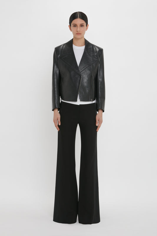 A person stands against a white background wearing a Victoria Beckham Tailored Leather Biker Jacket In Black, complemented by a white undershirt and black wide-leg pants.