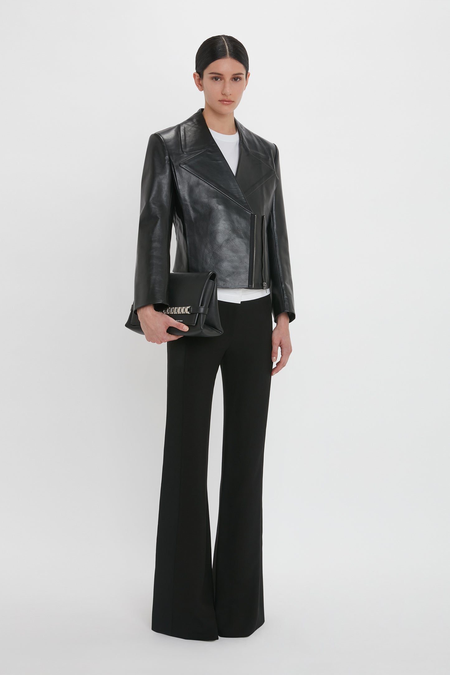 A person with dark hair is wearing a black Tailored Leather Biker Jacket In Black by Victoria Beckham made of calf leather, a white shirt, black flared pants, and holding a black handbag. They are standing against a plain white background.