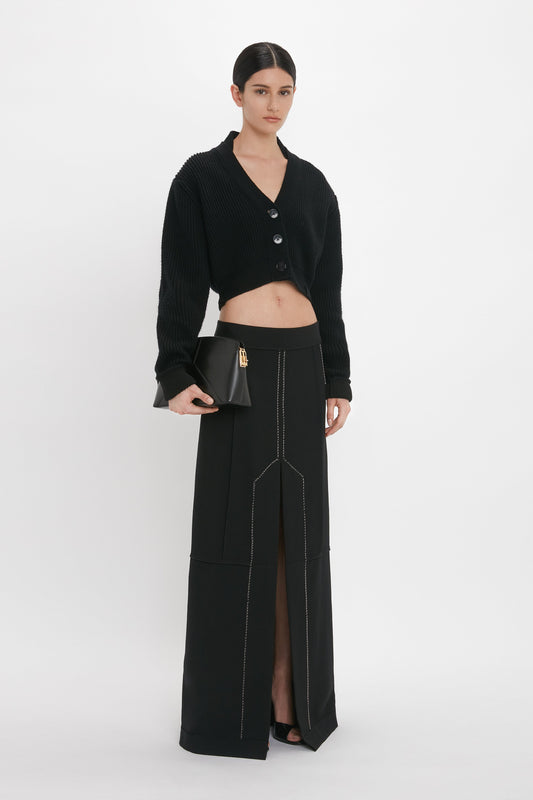 A woman stands against a plain background wearing a black cropped cardigan, a Deconstructed Floor-Length Skirt In Black by Victoria Beckham with a front slit, and holding a black clutch purse.