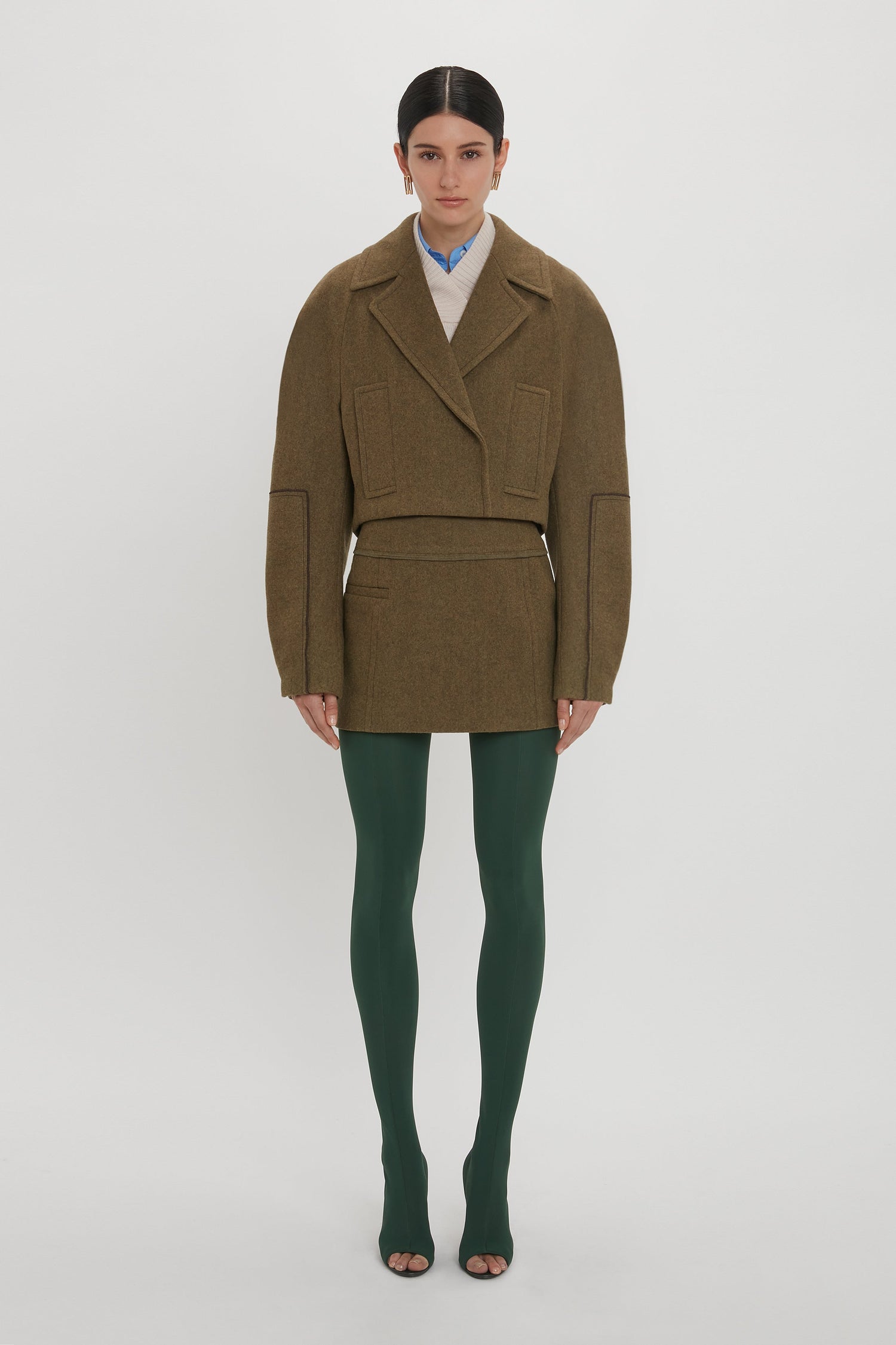 Person standing against a plain background, wearing an oversized brown jacket, green tights, and open-toe shoes. The outfit is complemented by a chic Victoria Beckham Tailored Mini Skirt In Khaki, adding a refined touch to the overall casual look.