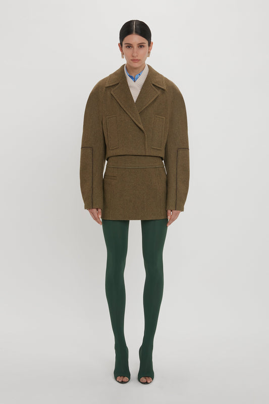 Person standing against a plain background, wearing an oversized brown jacket, green tights, and open-toe shoes. The outfit is complemented by a chic Victoria Beckham Tailored Mini Skirt In Khaki, adding a refined touch to the overall casual look.