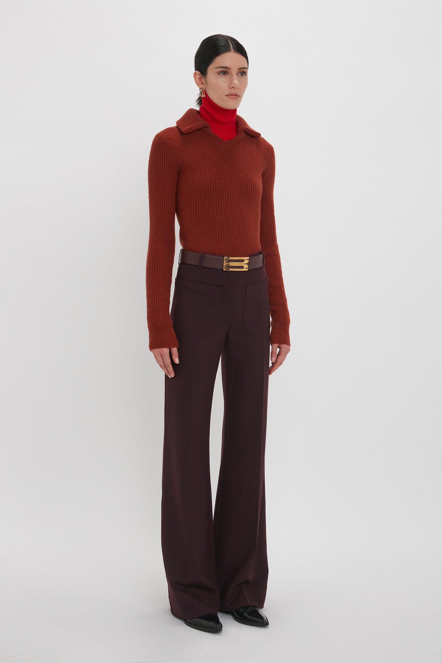 A person stands against a plain background wearing a Victoria Beckham Double Collared Jumper In Russet, a red turtleneck underneath, maroon trousers, a brown belt, and black shoes.