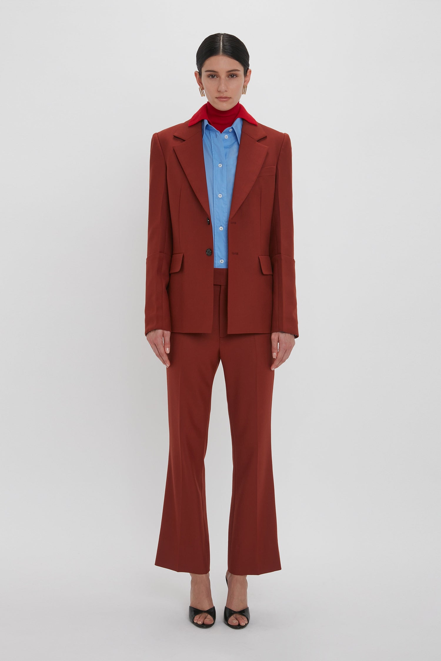 A person stands against a plain white background, wearing a rust-colored suit over a light blue button-up shirt and Wide Cropped Flare Trouser In Russet by Victoria Beckham, complemented by a red scarf around the neck and black open-toe heels.