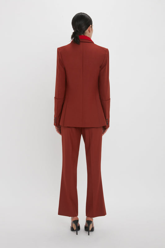 A person with black hair is shown from the back, wearing a red tailored suit with 1970s-inspired Wide Cropped Flare Trouser In Russet and black high-heeled shoes, standing against a plain white background—a nod to Victoria Beckham’s highlights for AW24.